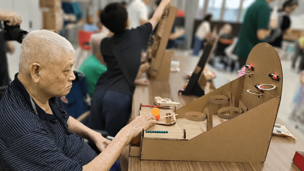 An elderly man sits at a table, focused on playing with a handmade cardboard game featuring ramps, rings, and an orange ball, while a busy activity room with other participants is softly blurred in the background.